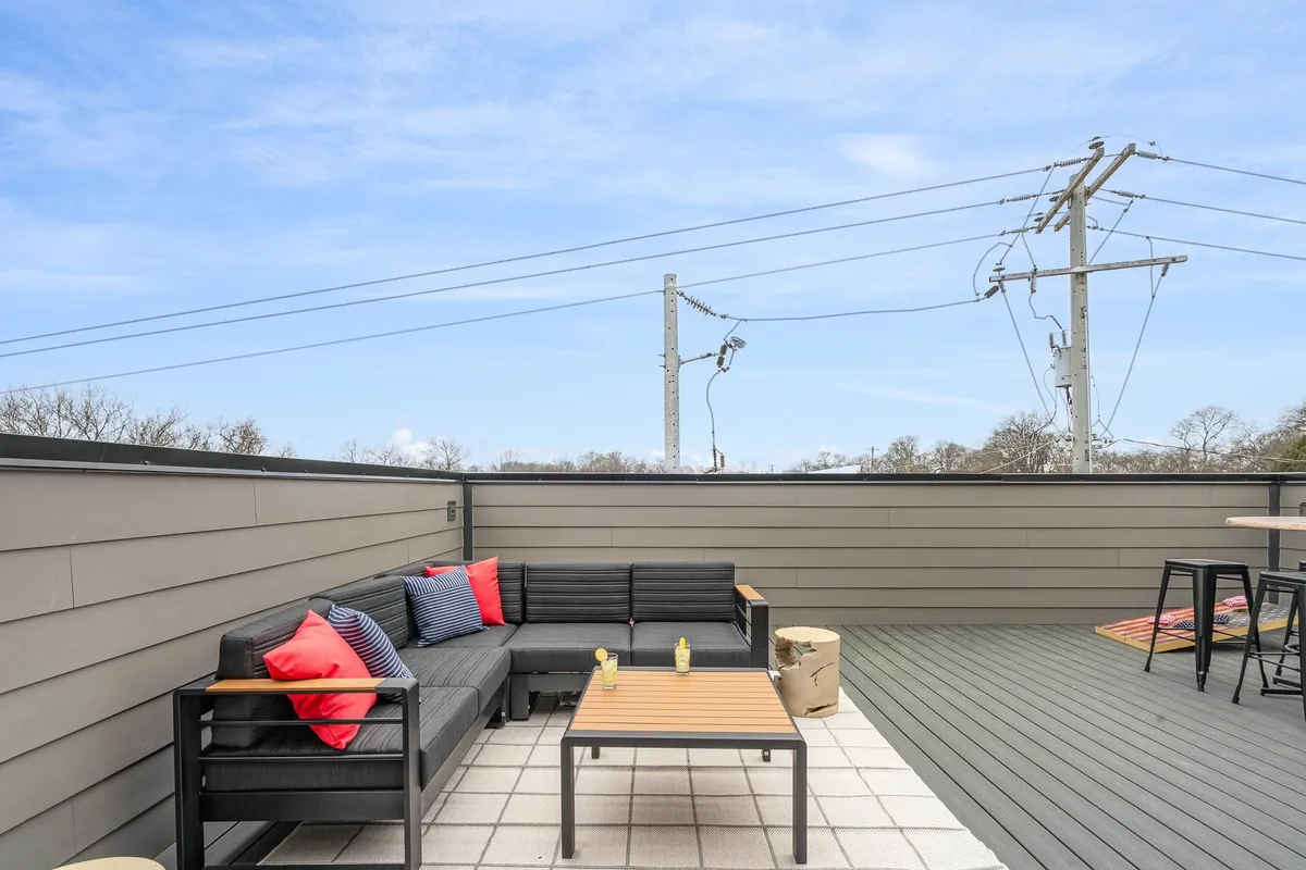 A cozy exterior seating area is arranged with decorative cushions and a table topped with refreshments, set against an open sky backdrop. The scene is intimate and welcoming, conveying a perfect spot for socializing, dining al fresco, or enjoying drinks while taking in skyline views.