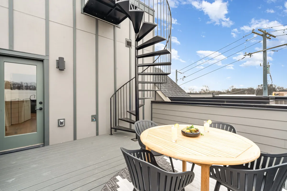 A stylish rooftop patio is pictured, featuring a spiral staircase that leads up to a level with a dining table surrounded by outdoor chairs. The scene includes ample open space, ideal for social gatherings, and the city skyline is visible in the background, hinting at elevated views. The setup offers an inviting space for dining, relaxing, or entertaining.