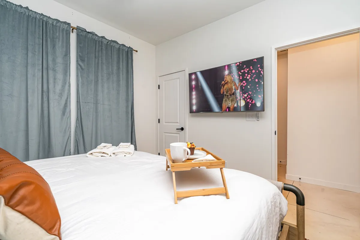 A cozy bedroom with crisp white linens on the bed, a Smart TV displaying a live performance, a breakfast tray, and soft drapes. The overall mood is inviting, perfect for unwinding after a day of exploring.