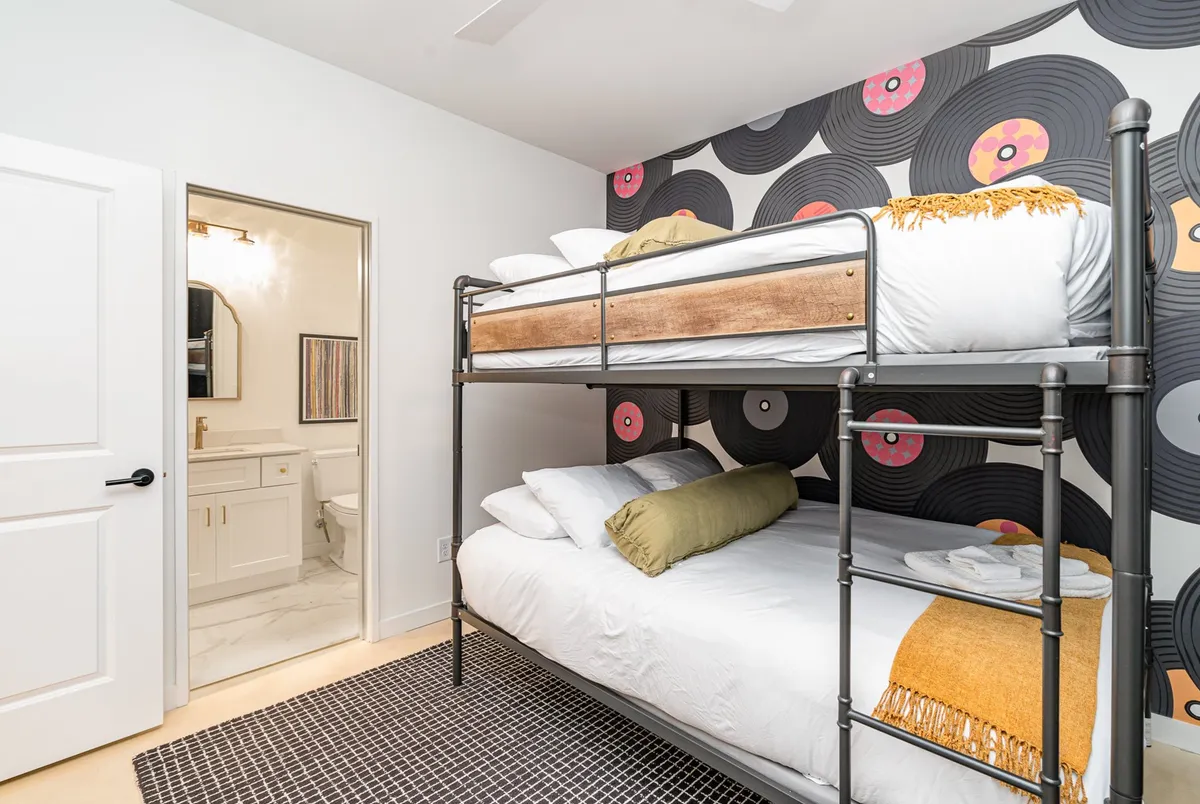 A playful bunk room featuring queen-sized beds stacked in a bunk setup, with colorful vinyl record wallpaper adding character. There's a visible bathroom door, making the connection to en-suite or shared bath convenience clear.