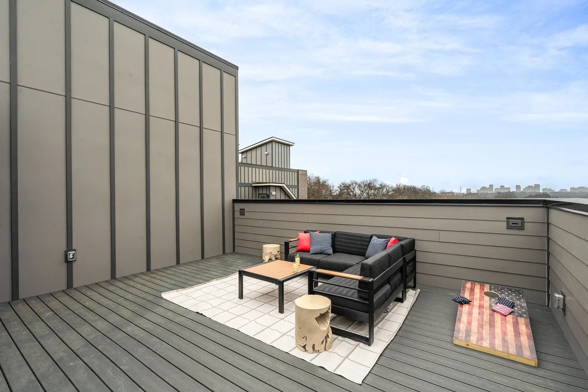 A stylish rooftop patio features an inviting outdoor seating area with comfortable chairs, a coffee table, and decorative pillows. The space is adorned with American flag-themed decor and is bathed in warm natural light, suggesting an atmosphere perfect for social gatherings or sunset relaxation.