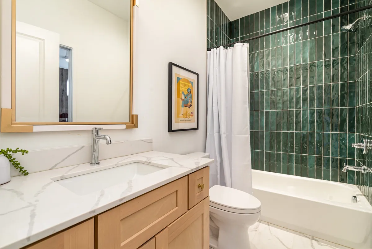 An elegant bathroom with a marble countertop, green tiled walls, gold-framed mirror, and decorative artwork. The mood is serene and inviting, offering a glimpse of the high-end finishes and peaceful atmosphere.