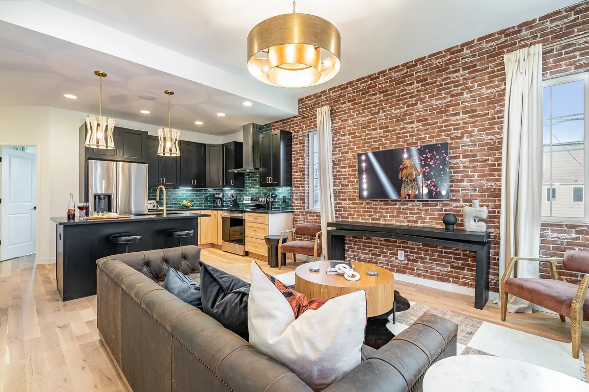 The image showcases a spacious, modern living area with a plush sectional sofa, a stylish brick accent wall, a large TV for entertainment, and a sleek kitchen island—creating a warm, inviting space perfect for gathering.