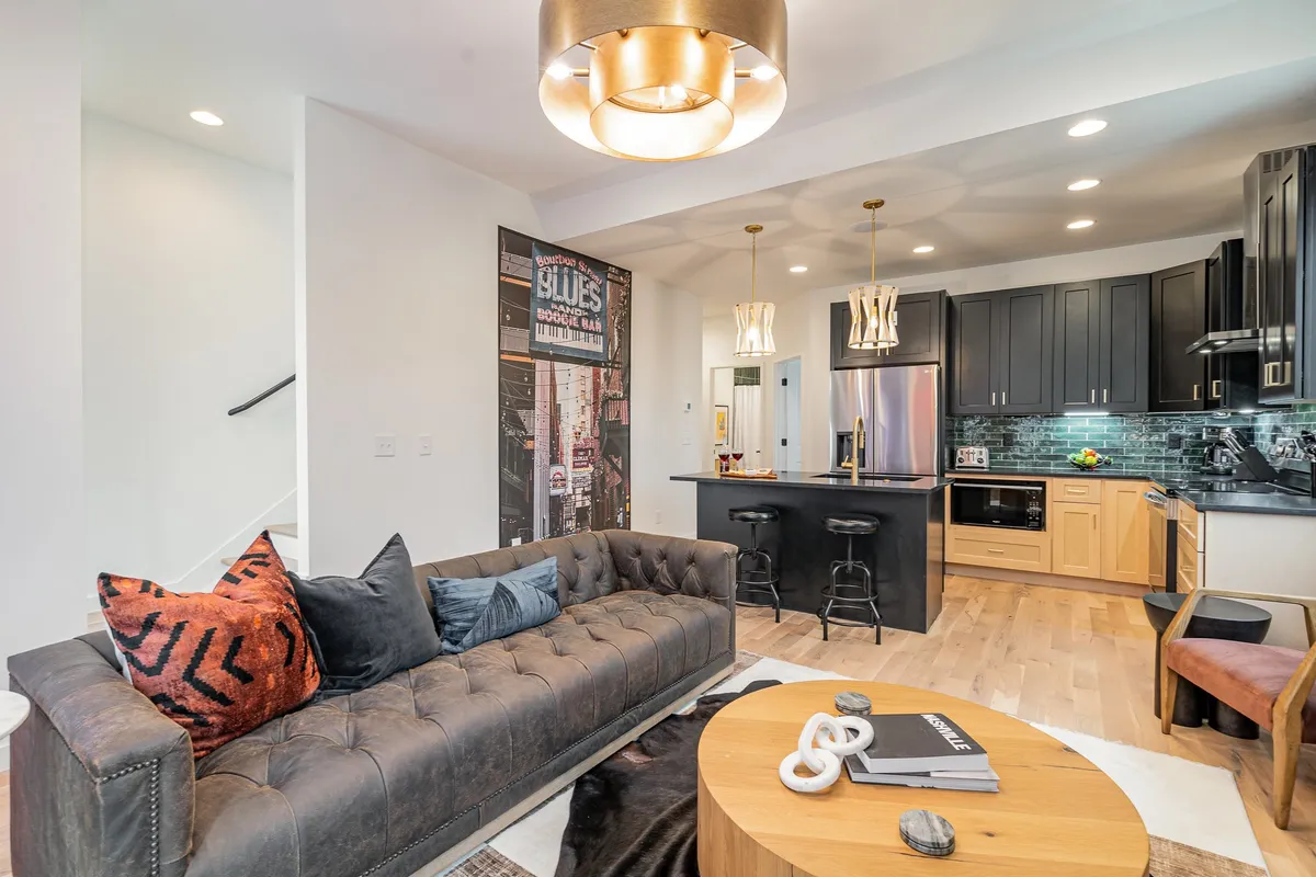 A cozy yet sophisticated living area combining a sofa, kitchen island with bar stools, decorative wall art, and modern lighting. The layout supports multiple activities from socializing to cooking in a warm, inviting ambiance.