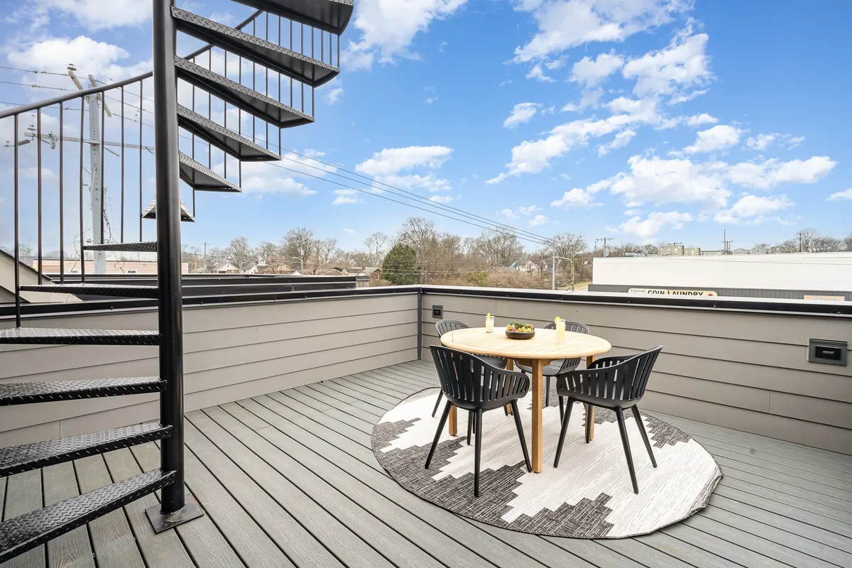 The image shows a relaxed rooftop setting with a round dining table surrounded by black outdoor chairs and a decorative rug. A spiral staircase connects different levels, reinforcing the multi-level aspect of the rooftop experience. Evening light casts a welcoming glow, evoking the mood of sunset festivities or late-night storytelling.