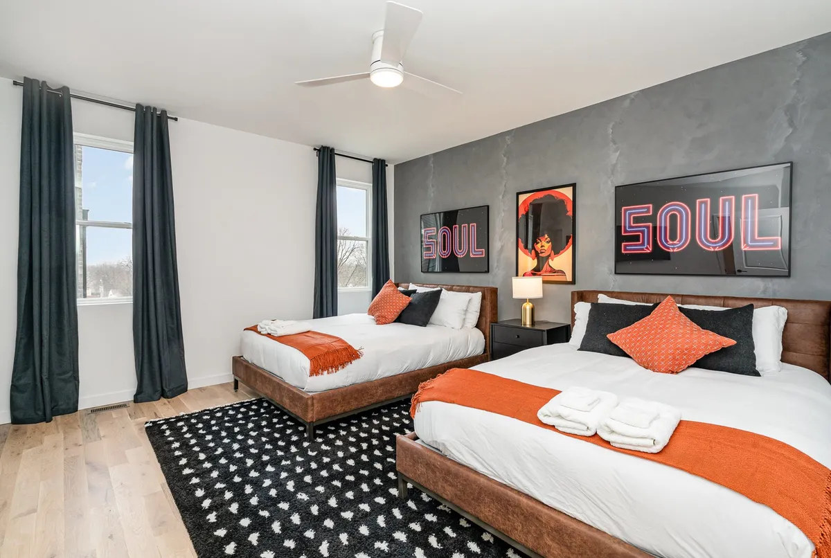 A stylish bedroom featuring two beds dressed in plush bedding, accented by eye-catching neon 'SOUL' artwork above. The room includes a black and white patterned rug and soft, ambient lighting, creating a relaxing yet artistic atmosphere ideal for both comfort and group stays.