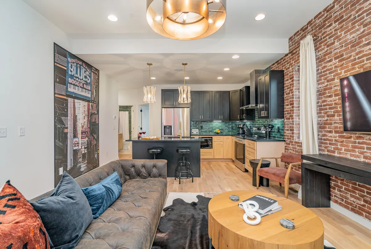 A contemporary open-plan space blending a chic sectional sofa, a modern kitchen island with seating, exposed brick wall, stylish light fixtures, and an artistic coffee table. The kitchen and living area are visibly integrated for seamless entertainment and socializing.